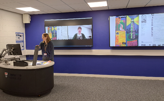Image of a woman standing at a desk (left of image) with an item from Special Collections under a visualiser and then to large screens on the wall (right of image) showing a Zoom call with Special Collections and the Special Collections items on the screen